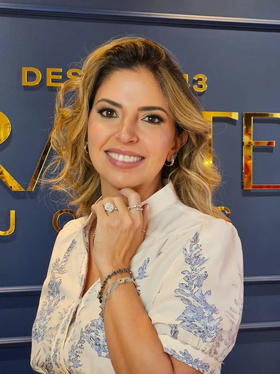 Mulher sorrindo e usando joias Arantes diante do fundo azul institucional da marca